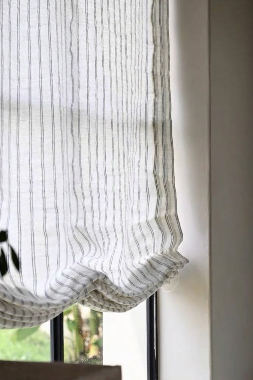 Roman shade in sheer white linen with narrow gray vertical stripes and softly gathered folds, shown lowered; gray striped roman shades