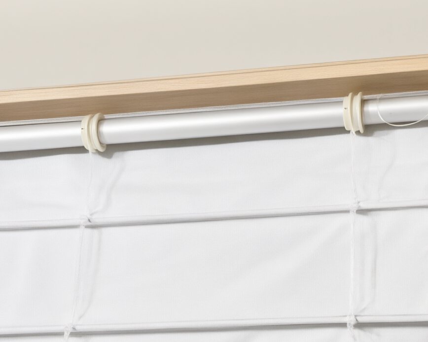 Close-up of a white pleated fabric blind or curtain panel hanging from a silver rod with cream-colored rings and a light wood header.
